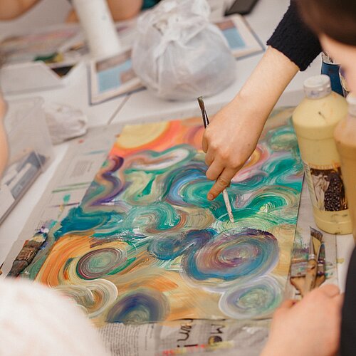 Mehrere Kinder malen mit Acrylfarbe ein Motiv aus bunten Kreisen auf ein Papier in der Mitte