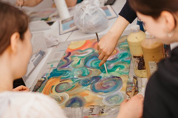 Mehrere Kinder malen mit Acrylfarbe ein Motiv aus bunten Kreisen auf ein Papier in der Mitte