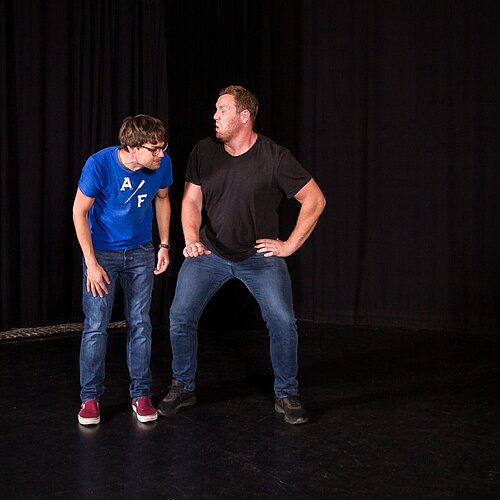 Zwei Improspieler stellen eine Szene dar vor schwarzem Bühnenhintergrund