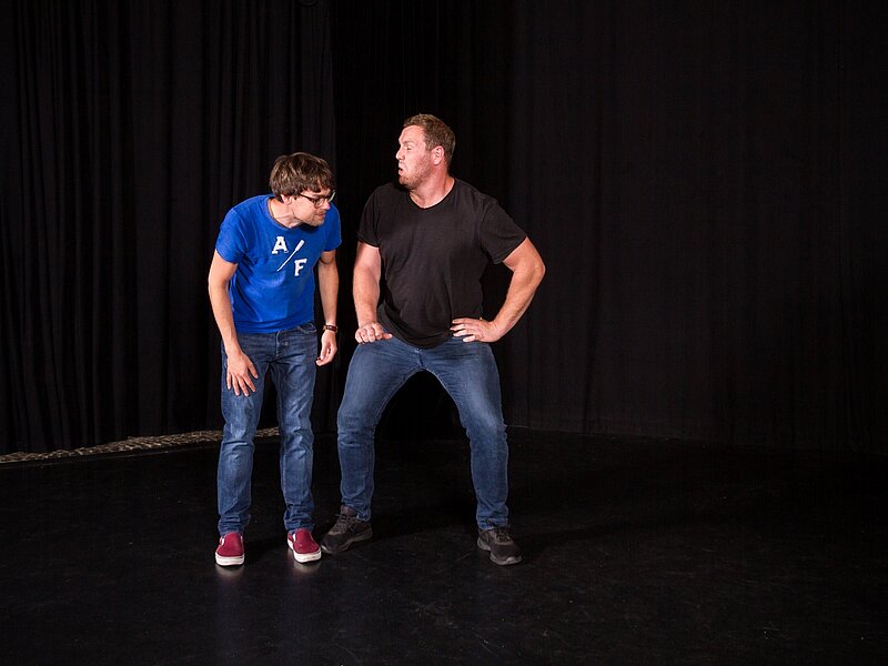 Zwei Improspieler stellen eine Szene dar vor schwarzem Bühnenhintergrund