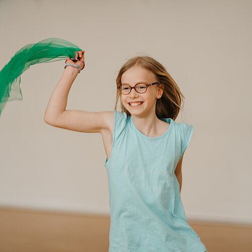 Maedchen schwingt froehlich ein leichtes gruenes Tuch im HIntergrund Tanzsaal