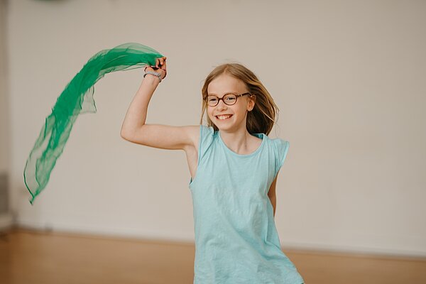 Maedchen schwingt froehlich ein leichtes gruenes Tuch im HIntergrund Tanzsaal