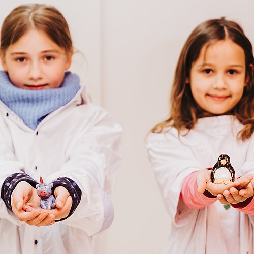 Zwei Maedchen in Werkkitteln zeigen stolz ihre getoepferten Figuren