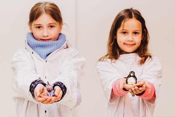 Zwei Maedchen in Werkkitteln zeigen stolz ihre getoepferten Figuren