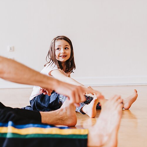 Kind und Tanzlehrer sitzen auf dem Boden und Dehnen sich in dem sie die bei gestrecktem Bein die Zehen mit den Haenden zum Oberkoerper ziehen.