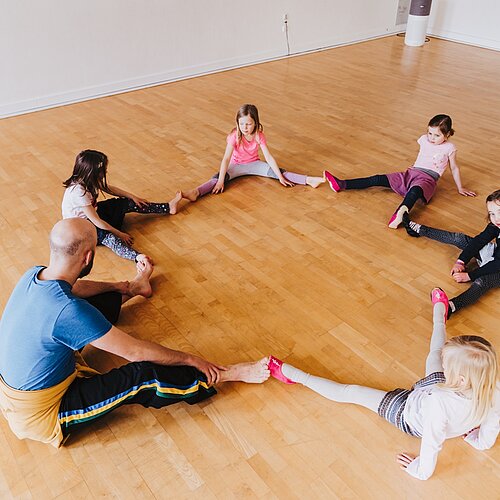 Kinder und Tanzlehrer sitzen mit gestreckten gespreizten Beinen im Kreis die Fuesse beruehren sich Ansicht von seitlich oben