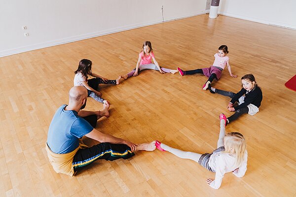 Kinder und Tanzlehrer sitzen mit gestreckten gespreizten Beinen im Kreis die Fuesse beruehren sich Ansicht von seitlich oben