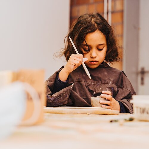 Maedchen an Werktisch bearbeitet konzentriert ein getaopertes Gefaess mit einem Werkzeug