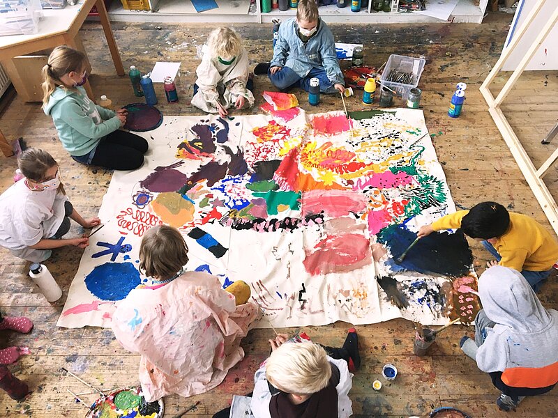 Eine Gruppe Kinder bemalen ein grosses weisses Tuch in ihrer Mitte mit bunter Farbe