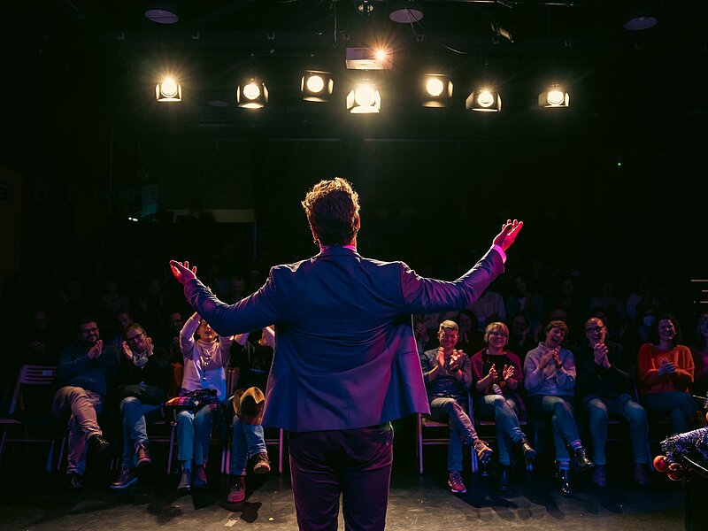 Rückenperspektive eines Künstler auf der Bühne des Kreativ-Hauses vor applaudierendem Publikum